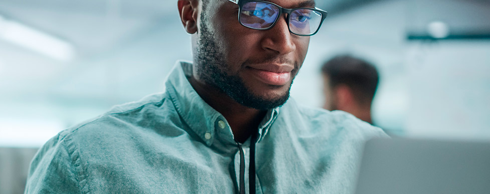 Homem negro, com barba e óculos, vestindo camisa verde, está olhando para baixo, como se estivesse lendo algo em uma tela, pois vemos o reflexo nas lentes de seu óculos.