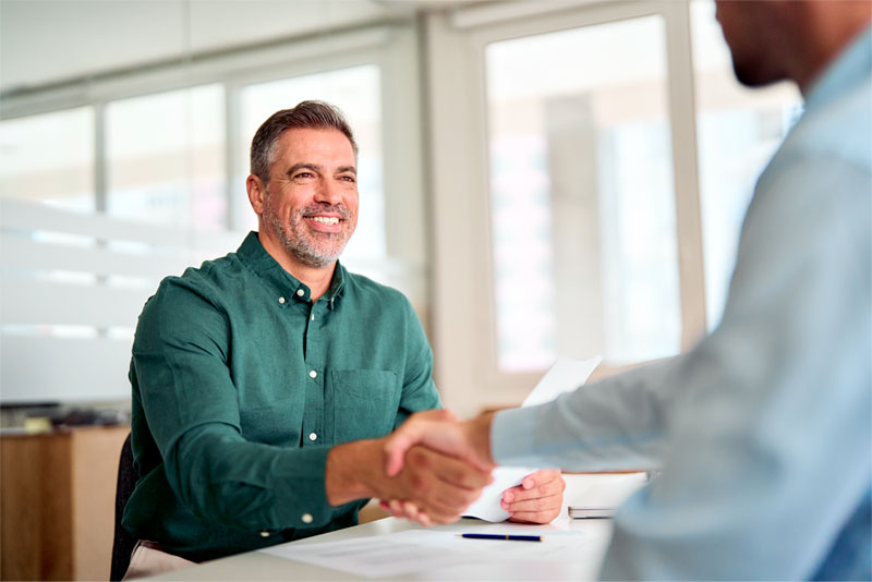 Homem branco com barba e de camisa verde está cumprimentando outro homem, que está um pouco desfocado na imagem, com um aperto de mão. O homem desfocado veste uma camisa branca. O cenário é um escritório. Ao fundo da imagem, é possível ver grandes janelas brancas.